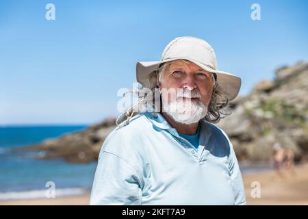 Uomo anziano dai capelli grigi bearded in cappello e t shirt blu in piedi sulla riva del mare godendosi il tempo soleggiato guardando lontano Foto Stock