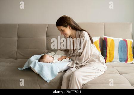 Giovane mamma in sleepwear toccare la mano del bambino mentre si siede sul divano nel fine settimana mattina nel soggiorno a casa Foto Stock