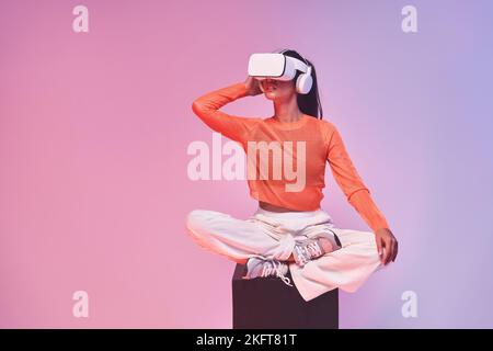 Giovane donna in un elegante vestito che tocca il visore VR e guarda lontano mentre siede a gambe incrociate sul cubo ed esplora il cyberspazio contro il backgrou viola Foto Stock