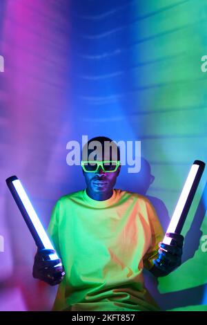 Maschio nero in occhiali da sole e abiti alla moda che tengono lampade fluorescenti mentre si siede in studio moderno con illuminazione al neon viola Foto Stock
