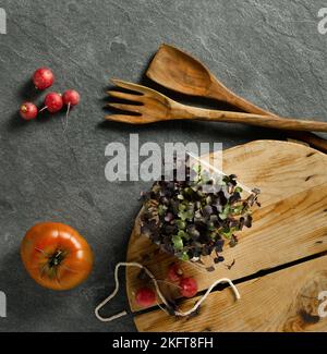 Vista dall'alto di pomodoro fresco e ravanelli sul tavolo con germogli verdi sul tagliere vicino a spatola e forchetta di legno Foto Stock
