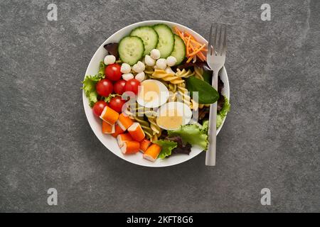 Insalata sana con verdure assortite e pasta con uova sode e verdure servite in ciotola con forchetta su tavolo di pietra Foto Stock