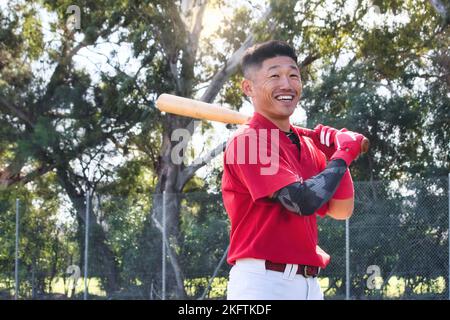 Un giovane asiatico che tiene una mazza da baseball in piedi in un parco da ballo sorridente Foto Stock