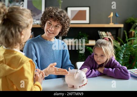 Madre che indica la banca del piggy e che insegna ai suoi bambini a risparmiare soldi mentre si siedono al tavolo nella stanza Foto Stock