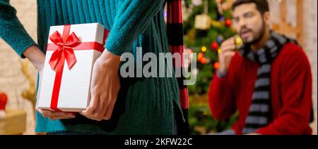 Vista posteriore giovane donna caucasica in pelle presenta per il ragazzo a sorpresa in celebrazione Natale a casa, donna in possesso di un regalo nascondere dietro per dare l'uomo w Foto Stock