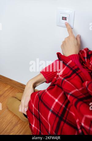 Primo piano della mano di una ragazza in un plaid caldo e calzini di lana trasforma il termostato di riscaldamento del pavimento ad una temperatura elevata. Impostazione della temperatura a casa Foto Stock