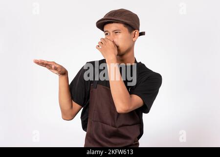 Bel barista asiatico con grembiule marrone e t-shirt nera isolata su sfondo bianco che copre il naso che odora qualcosa di puzzolente e disgustoso Foto Stock