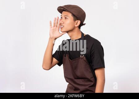 bel barista asiatico con grembiule marrone e t-shirt nera isolata su sfondo bianco che sussurra dicendo segreto Foto Stock