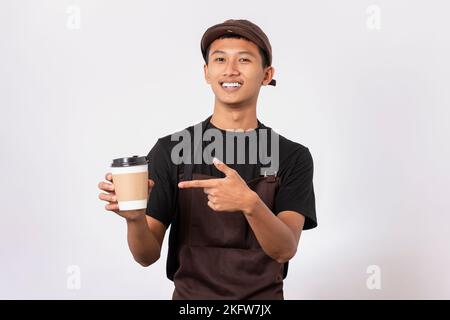 Bel barista asiatico con grembiule marrone e t-shirt nera isolata su sfondo bianco. Barista che tiene una tazza di caffè e serve il caffè per la crema Foto Stock
