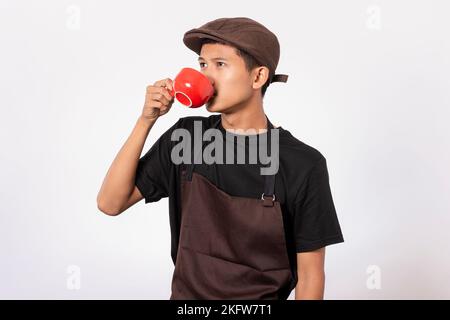 Bel barista asiatico con grembiule marrone e t-shirt nera isolata su sfondo bianco. Barista che tiene una tazza di caffè e serve il caffè per la crema Foto Stock