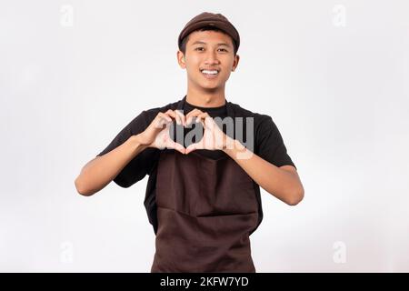 Bel barista asiatico con grembiule marrone e t-shirt nera isolata su sfondo bianco sorridente facendo forma simbolo del cuore con le mani. Foto Stock