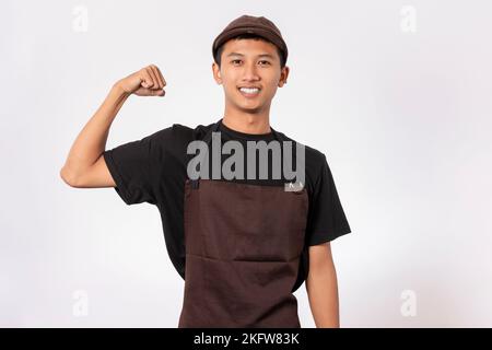 Bel barista asiatico con grembiule marrone e t-shirt nera isolata su sfondo bianco che mostra il bicep muscolare, la forma e il concetto sano Foto Stock