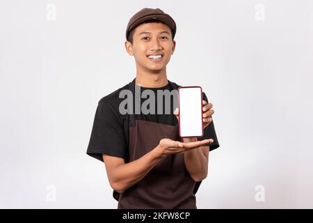 Bel barista asiatico con grembiule marrone e t-shirt nera isolata su sfondo bianco che tiene lo smartphone con schermo bianco per il mockup. Foto Stock