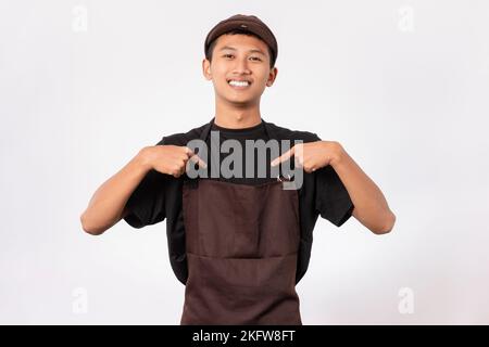 Bel barista asiatico con grembiule marrone e t-shirt nera isolata su sfondo bianco che indica te stesso con le dita Foto Stock