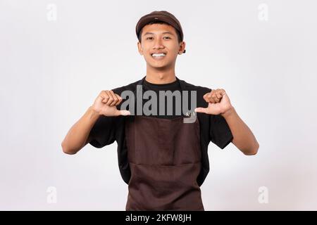 Bel barista asiatico con grembiule marrone e t-shirt nera isolata su sfondo bianco che indica te stesso con le dita Foto Stock