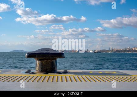 Paletto di ormeggio in metallo nero nella marina e nel terminal portuale. Paesaggio urbano di Noray sullo sfondo Foto Stock