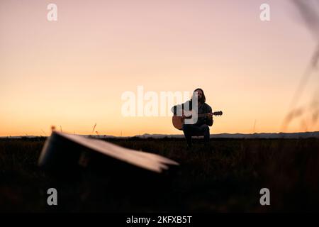 Giovane caucasico con barba seduta su uno sgabello concentrato con gli occhi chiusi suonando la chitarra al tramonto in un luogo solitario Foto Stock