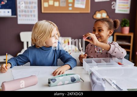 Allievo carino della scuola primaria usando l'affilatore della matita mentre si siede dalla scrivania vicino al ragazzo adorabile della scuola bionda che la guarda alla lezione Foto Stock