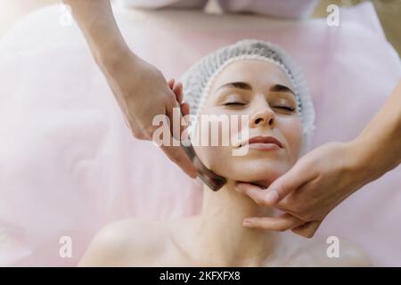 Massaggio con una speciale paletta massaggiante, grilletto per impastare sul viso, primo piano. Massaggio lama di una donna europea. Vista dall'alto Foto Stock