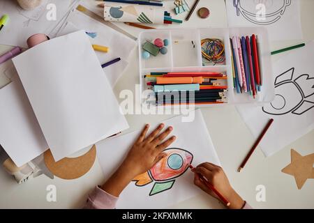 Vista dall'alto delle mani del piccolo studente afro-americano della scuola materna che disegna un razzo con pastelli multicolore sulla carta a lezione Foto Stock