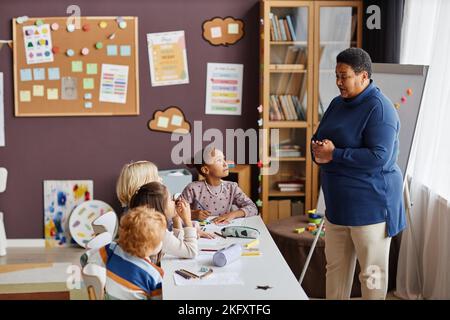 Insegnante fiducioso maturo in casualwear fare la presentazione a simpatici studenti interculturali della scuola primaria a lezione di disegno Foto Stock