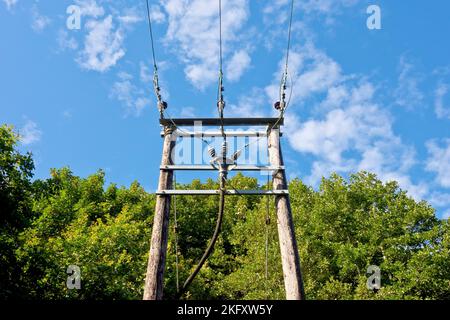 Un robusto pilone di elettricità rurale in legno costruito per trasportare tre cavi ad alta tensione attraverso un fiume, sparato contro un cielo blu nuvoloso. Foto Stock