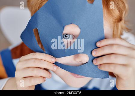 Primo piano di un simpatico ragazzino blueeyed che copre il viso con una maschera di carta blu fatta a mano, mantenendo occhio e naso nei fori tagliati Foto Stock