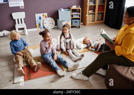 Gruppo di curiosi ragazzi interculturali che si rilassano sul tappeto davanti all'insegnante che legge loro favola nel miidle del giorno alla scuola materna Foto Stock