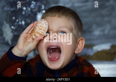 Ritratto di simpatico bambino allegro. Il ragazzo gioca con i tangerini tra le decorazioni natalizie. Atmosfera festosa per la vigilia di Capodanno. Elegante e accogliente Foto Stock