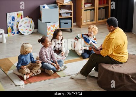 Diversi adorabili ragazzi interculturali si sono riuniti sul pavimento di fronte al loro insegnante di scuola materna leggendo loro un libro di racconti Foto Stock