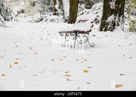 Panchina del parco coperta di neve nel tranquillo parco cittadino. Foglie gialle cadute sulla neve. Focalizzazione selettiva sul banco Foto Stock