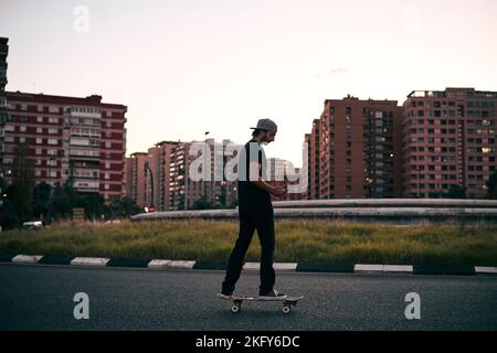 Giovane caucasico in una t-shirt nera e pantaloni neri che cavalcano pericolosamente su uno skateboard mentre manipola lo schermo del suo smartphone in città Foto Stock