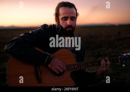 giovane uomo caucasico con i capelli raccolti seduto in mezzo a un grande campo erboso calma e concentrato suonare la chitarra appassionatamente al tramonto Foto Stock