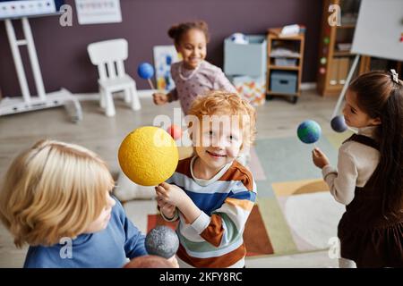 Simpatico allievo della scuola primaria sorridente con il modello di sole che guarda la macchina fotografica mentre si trova in piedi tra gli altri bambini che giocano con pianeti in asilo Foto Stock