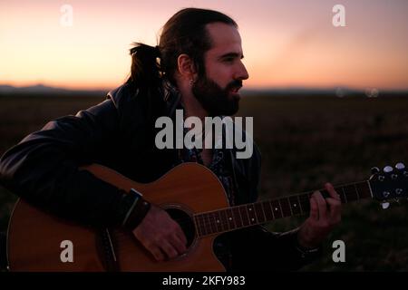 Giovane caucasico con una barba in profilo seduta in mezzo a un grande campo erboso calma e concentrata suonare la chitarra al tramonto in un Foto Stock