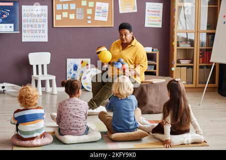 Insegnante di scuola materna seduto di fronte a bambini interculturali e parlando con loro di pianeti del sistema solare a lezione in classe Foto Stock