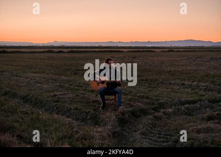 Giovane caucasico con barba e capelli lunghi seduto su uno sgabello serenamente suonando la chitarra al tramonto in un posto solitario Foto Stock