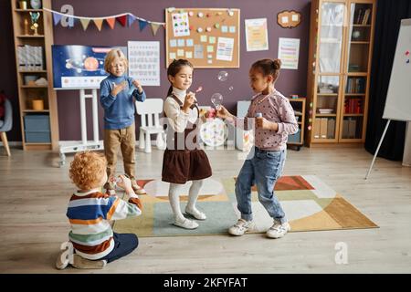 Gruppo di bambini piccoli spensierati in casualwear divertirsi con le bolle di sapone mentre in piedi sul tappeto in classe della scuola materna Foto Stock