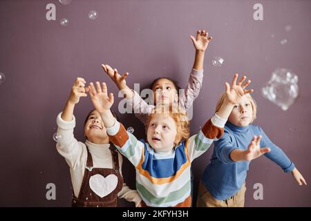 Gruppo di allegri bambini carino in casualwear alzando le mani mentre cattura bolle di sapone contro la parete nella scuola materna Foto Stock