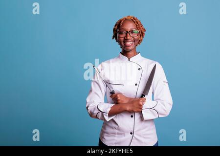 Chef afroamericano in uniforme sorridente guardando la macchina fotografica tenendo coltello e affilatore con le braccia incrociate. Cuoca femminile professionista con utensili da cucina in studio girato su sfondo blu. Foto Stock