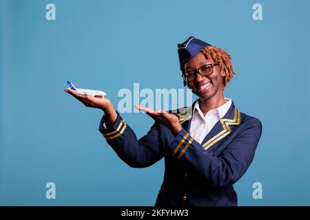 Sorridente afroamericano che indica l'aeroplano commerciale che tiene in mano per la pubblicità della linea aerea. Assistente di volo che indossa l'uniforme dei membri dell'equipaggio nella foto in studio. Foto Stock