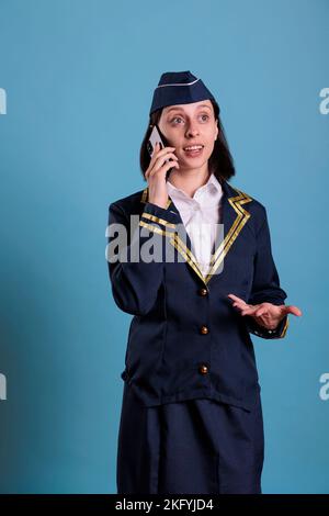 Sorrido che non abbia una chiamata remota tramite telefono cellulare. Assistente di volo in aviazione uniforme rispondere chiamata, hostess aereo in piedi con telefono in mano, guardando la macchina fotografica, scatto medio Foto Stock