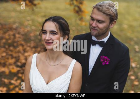 Foto romantica di matrimonio autunnale. Bella donna mediorientale sognante positiva in abito da sposa senza maniche in piedi di fronte al suo marito biondo scandinavo. Inquadratura media all'aperto. Foto di alta qualità Foto Stock