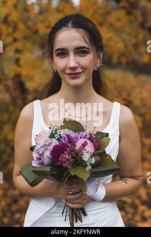 Stupendo verticale medio ritratto di bella sposa Medio Oriente in er fine 20s guardando la macchina fotografica, sorridendo dolcemente, e tenendo bello fresco fiori bouquet. Foglie d'autunno dorate sfocate sullo sfondo. Foto di alta qualità Foto Stock