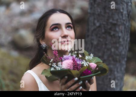 Concetto di fotoshoot nuziale. Bella sposa etnica in posa all'aperto nella foresta. Primo piano ritratto di attraente donna turca che guarda in su e tenendo il suo bouquet nuziale accanto al suo volto. Foto di alta qualità Foto Stock