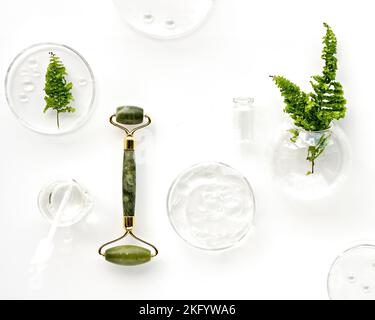 Moisturizer self-made e rullo verde di giada. Foglie di felce esotiche e gocce d'acqua su sfondo bianco. Massaggio viso, cosmetici fatti a mano Foto Stock