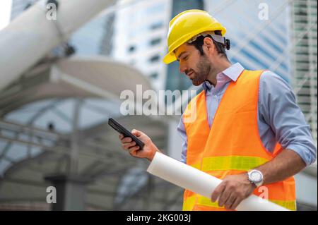 Gli ingegneri dell'uomo caucasici usano uno smartphone per parlare, indossando un giubbotto arancione e un grande elmetto, e l'altra mano che tiene la pianta bianca del sito Foto Stock