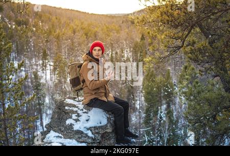 Un turista-viaggiatore con uno zaino in un cappello rosso siede sul bordo di una scogliera e guarda ad una valle di inverno della foresta. Il concetto di libertà e di unità con Foto Stock