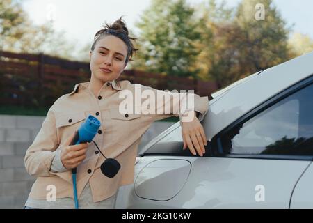 Giovane donna che tiene il cavo di alimentazione dalla sua auto elettrica, preparato per caricarlo in casa, sostenibile e concetto di trasporto economico. Foto Stock