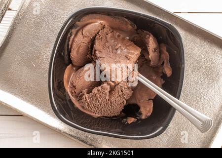 Gelato al cioccolato marrone scuro in una ciotola di ceramica nera con un cucchiaio di metallo su un vassoio di metallo, primo piano, vista dall'alto. Foto Stock